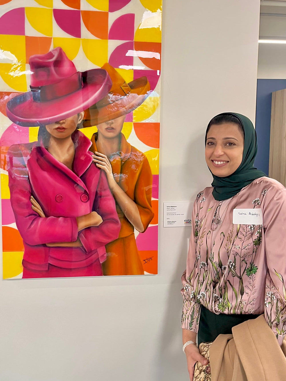 Visual artist Salma Abdelaziz  in a green hijab and pink patterned blouse stands beside a colorful fashion art print on a gallery wall. The print features two women in pink and orange clothing against a geometric yellow, pink, and orange background.